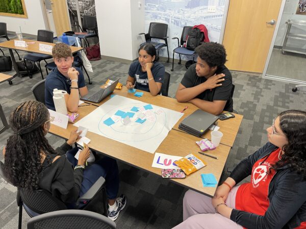 Students sit at a table. On the table is a piece of poster board with blue sticky notes.