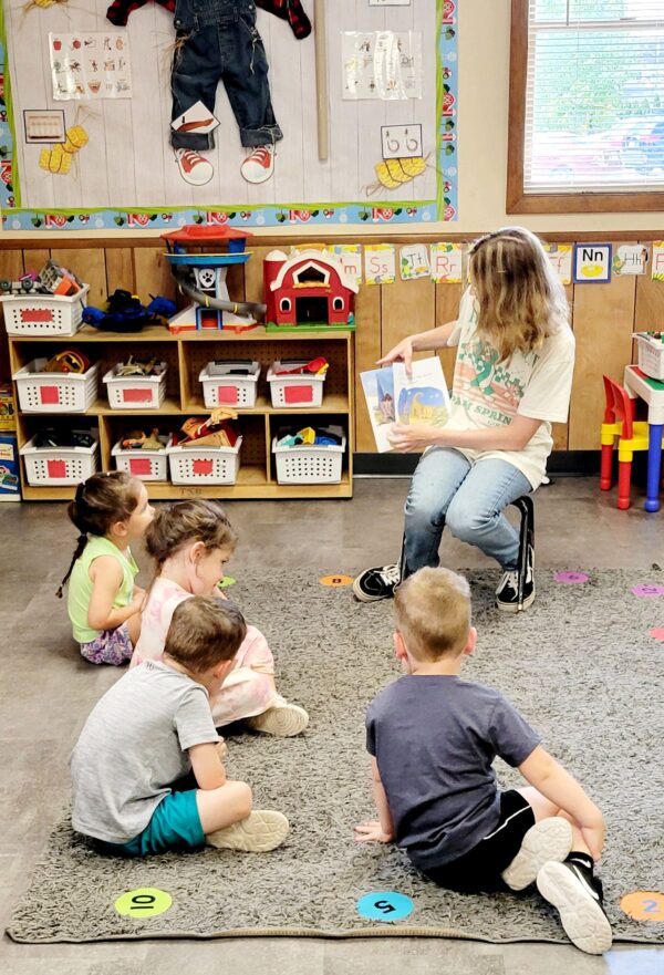 A teacher reads to a small group of young students who are gathered around her on a carpet