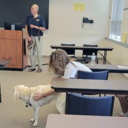 Service Paws President Joe Fagnani speaks at the front of a classroom while a student pets his dog, Phoebe, a yellow labrador retriever.