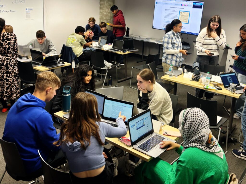 A group of students sit at tables and work on laptops in groups.