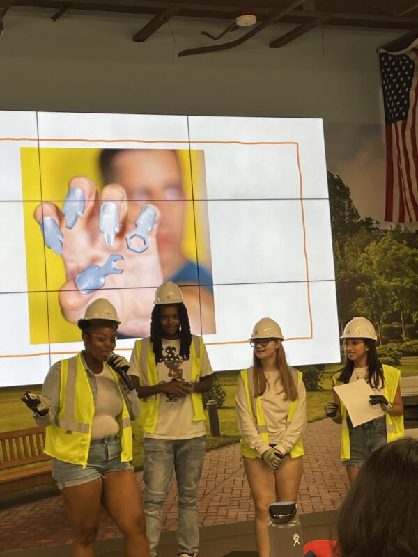 Students dressed in yellow and wearing yellow hard hats stand in front of a screen to give a presentation