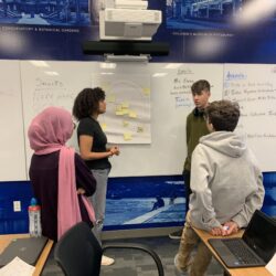 Four students have a discussion about information on a poster board in a classroom