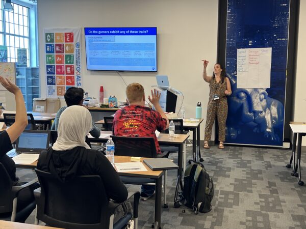 An instructor at the front of a classroom points at students who are raising their hands to ask questions.