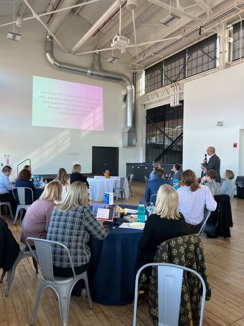 Attendees at the Readiness Institute Summit sit at tables and watch a presentation being projected onto the wall  