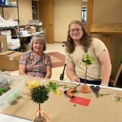 Two people sit at a table where they have been crafting model trees.