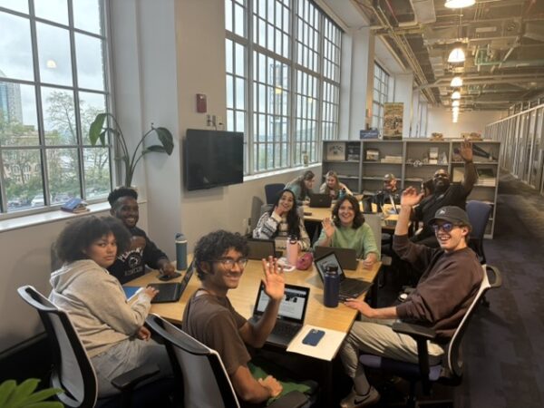 students at tables with computers, smiling and waving to camera