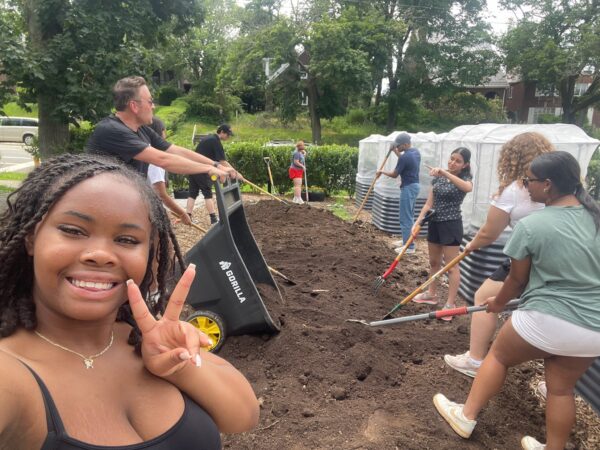 girl in forefront smiling and peace sign to camera while other students in background shovel soil at Sunny's Community Garden