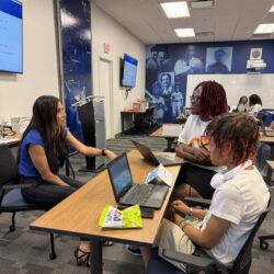 Students engage with an instructor while sitting at a table with laptops.