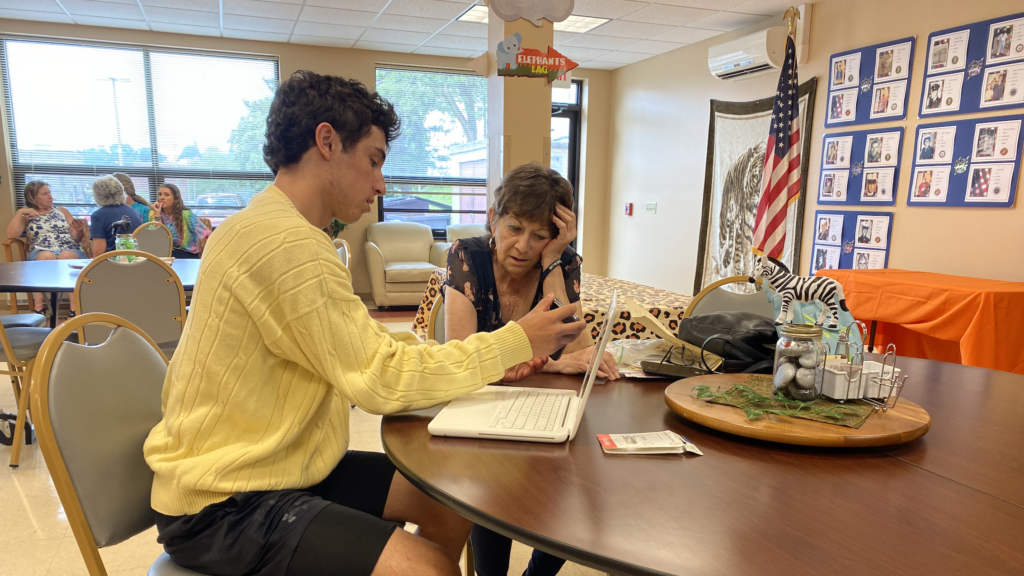 A Summer Program learner sits at a table and explains the use of a cell phone to a resident while volunteering with Blair Senior Services.